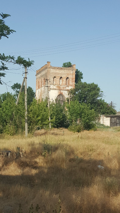 Старовинна прямокутна вежа з червоної цегли та арочними віконними прорізами, що виглядає з-поза зелених дерев і кущів. Споруда має ознаки занедбаності та великі дерев'яні ворота в нижній частині. На передньому плані — поле з сухою жовтою травою, електричні стовпи та дроти на тлі безхмарного блакитного неба.