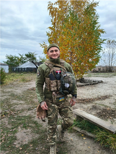 Усміхнений український військовий у камуфляжній формі, бронежилеті та з каскою в руках стоїть на тлі осіннього дерева із жовтим листям. 