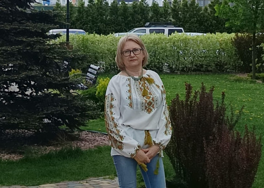 Жінка в окулярах та українській вишиванці й джинсах стоїть на галявині у сквері біля високої ялини. На задньому плані видно зелені насадження, декоративні ліхтарі та багатоповерхові будинки. 