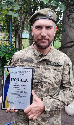 Портрет українського військовослужбовця (ветерана) у піксельній військовій формі та береті, який тримає в руках грамоту в рамці.