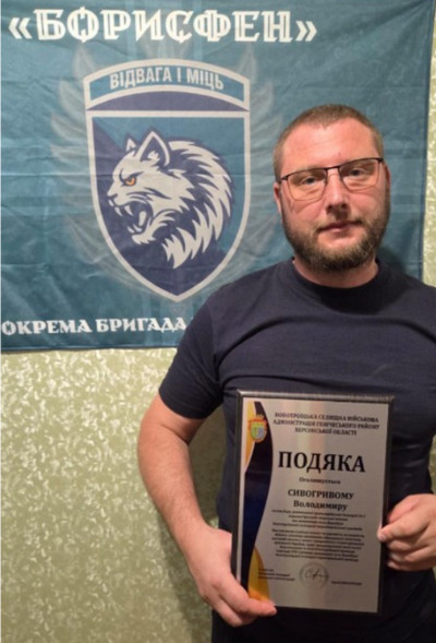 Чоловік у чорній футболці та окулярах тримає в руках рамку з грамотою «ПОДЯКА» від Новотроїцької селищної військової адміністрації Генічеського району Херсонської області, виданою Сивогривому Володимиру. На задньому плані висить темно-зелений прапор із написом «БОРИСФЕН», гаслом «ВІДВАГА І МІЦЬ» та гербом із зображенням рисі. 