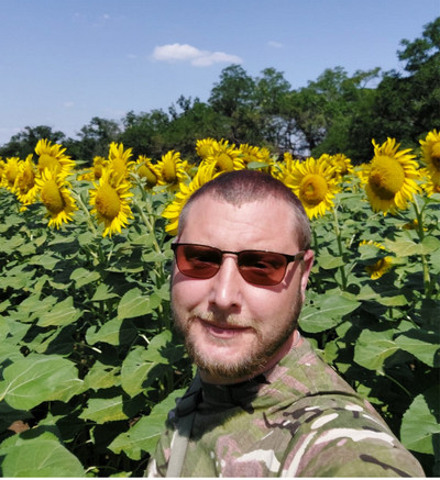 Чоловік у сонцезахисних окулярах та камуфляжному одязі робить селфі на тлі розквітлого поля соняшників. На задньому плані видно зелені дерева та блакитне небо з декількома легкими хмарами під яскравим сонячним світлом.