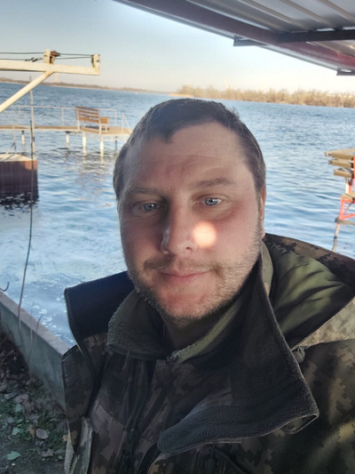 Чоловік у військово-камуфляжній куртці робить селфі на тлі річки з дерев'яним пірсом.