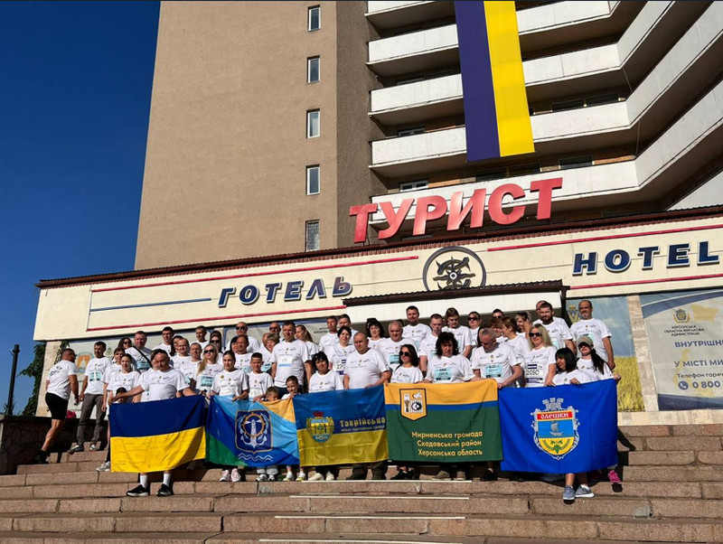 Група учасників патріотичного забігу у білих футболках стоїть на сходах перед готелем «Турист». Учасники тримають у руках Державний Прапор України та прапори громад Херсонщини. На фасаді будівлі розгорнуто великий синьо-жовтий стяг.