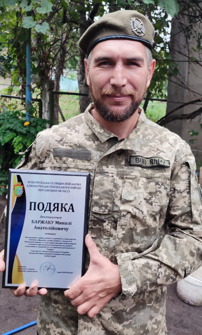 Військовослужбовець з бородою, у формі та береті, тримає грамоту «Подяка» від Новотроїцької військової адміністрації на ім'я Баржака Миколи Анатолійовича. У грамоті висловлюється вдячність за службу. Він стоїть на вулиці, на тлі зелених кущів.
